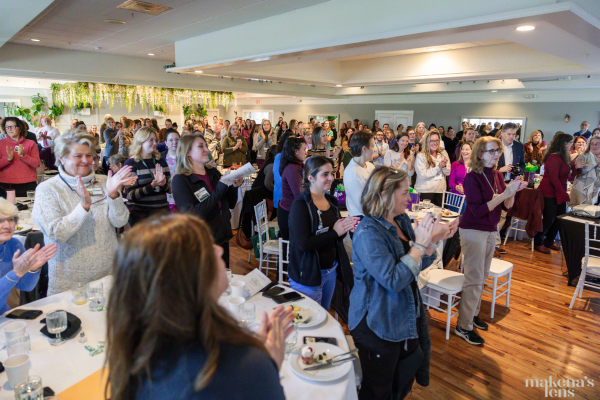 Large group of employees standing and applauding during an employee appreciation event at The Oaks Grandview venue. Attendees fill the bright, modern event space decorated with greenery, enjoying a celebratory gathering with tables set for a luncheon. Corporate appreciation event at The Oaks Grandview.