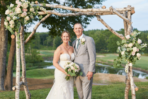 Couple standing beneath a rustic wooden ceremony arch on the lawn at The Oaks Grandview Venue, overlooking rolling greens and water, ideal for an intimate micro wedding ceremony.