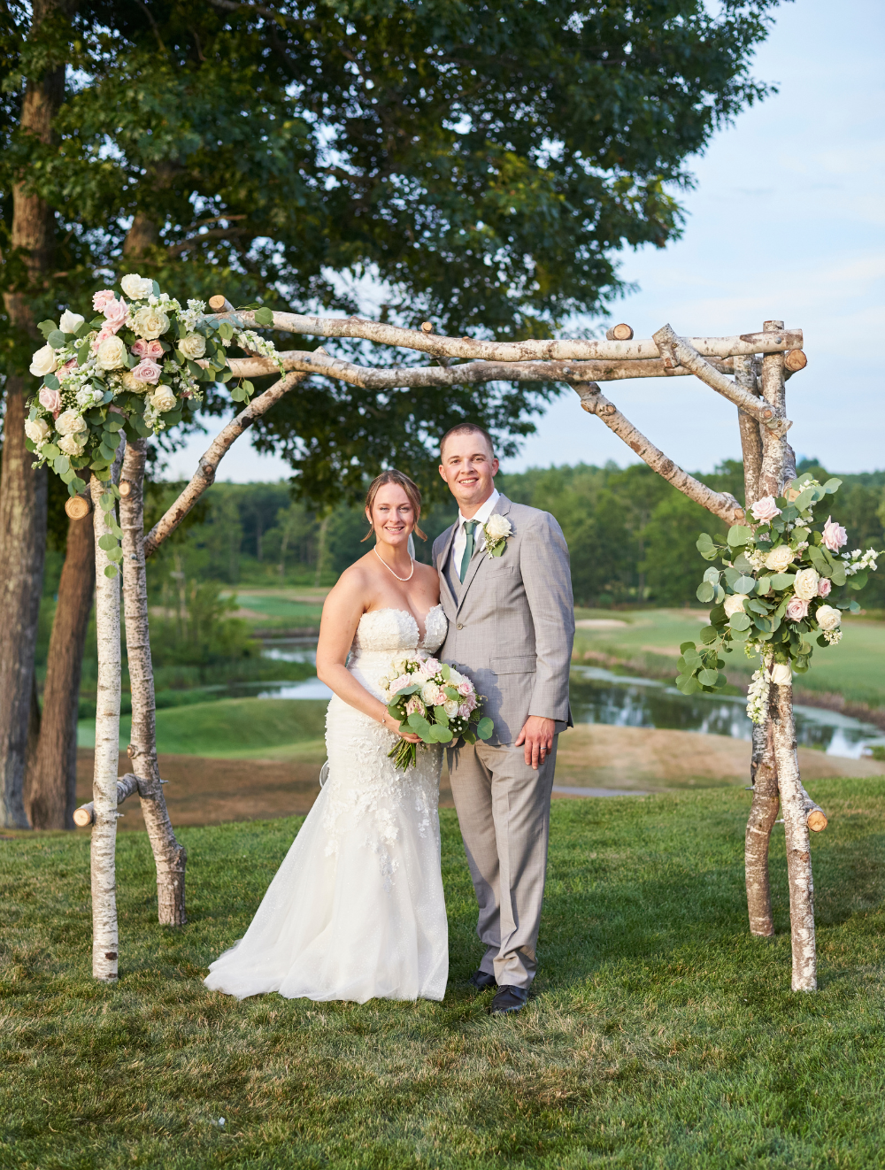 Couple standing beneath a rustic wooden ceremony arch on the lawn at The Oaks Grandview Venue, overlooking rolling greens and water, ideal for an intimate micro wedding ceremony.