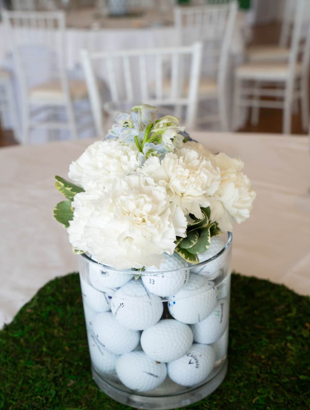 Centerpiece for the tables in the ballroom at The Grandview Venue. Golf balls fill up clear vases, topped with white flowers, on a grass patch mat.